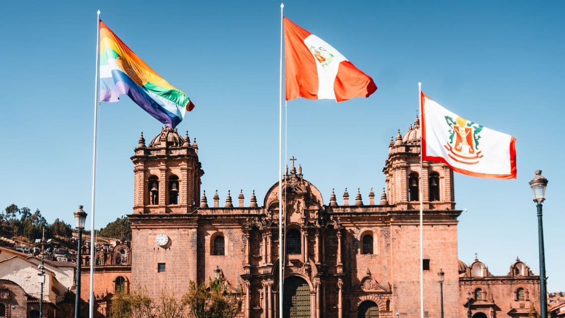 Three flags, including a rainbow flag, fly on flagpoles in front of a large, ornate stone cathedral under a clear blue sky.