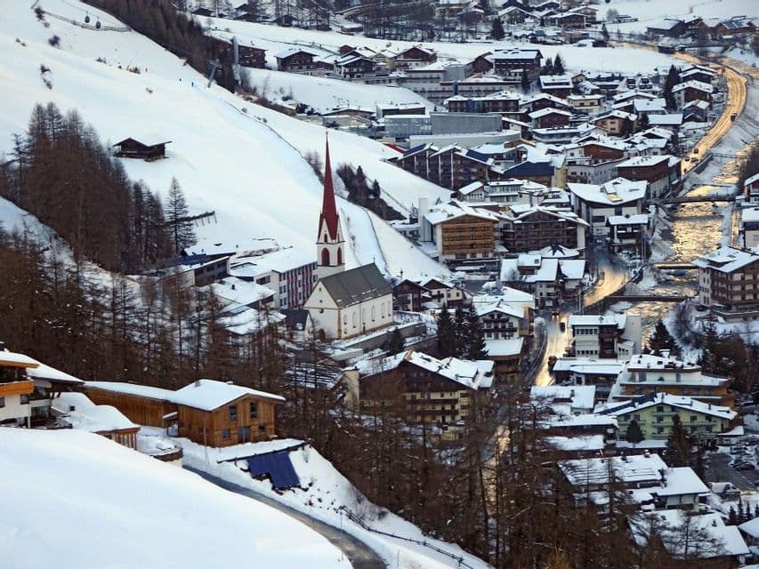 Ein alpines Dorf mit einer rot getürmten Kirche liegt in einem schneebedeckten Bergtal.