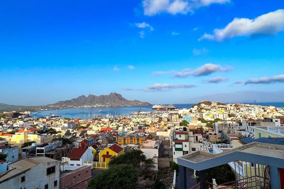 Ein Panoramablick auf eine farbenfrohe Küstenstadt mit einem Hafen voller Boote und Bergen auf der anderen Seite des Wassers unter blauem Himmel.