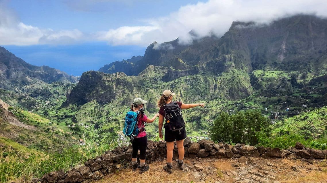 Zwei Frauen einer WeRoad-Gruppenreise stehen auf einem Felsvorsprung und zeigen auf ein üppiges grünes Bergtal mit dem Meer in der Ferne.