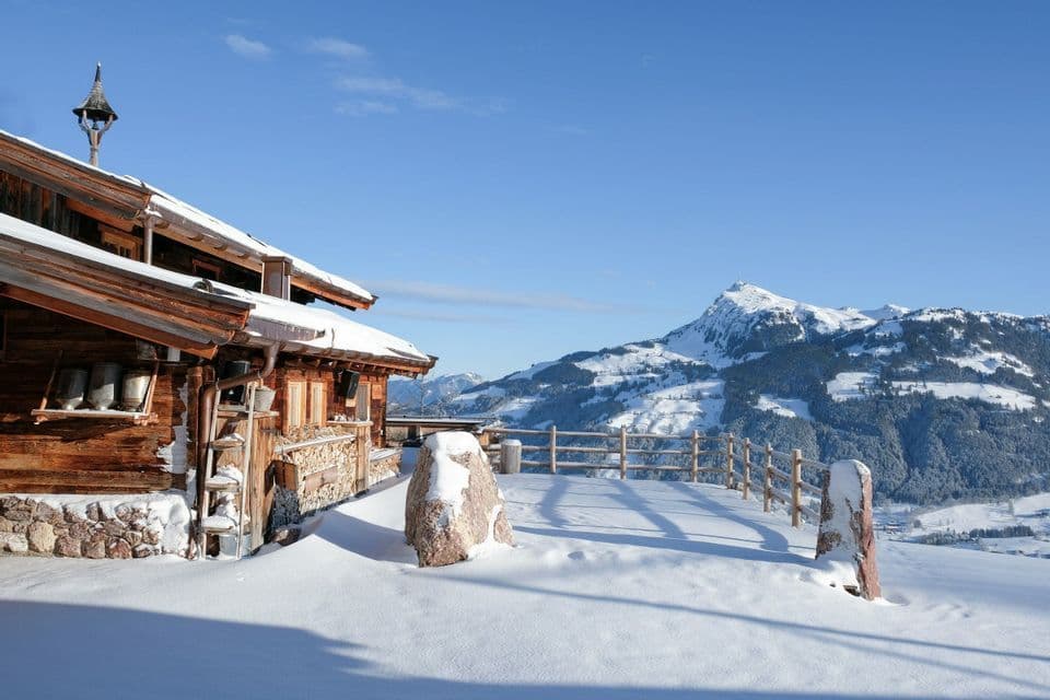 Un chalet en bois avec terrasse est recouvert de neige, surplombant une chaîne de montagnes enneigées sous un ciel bleu clair.