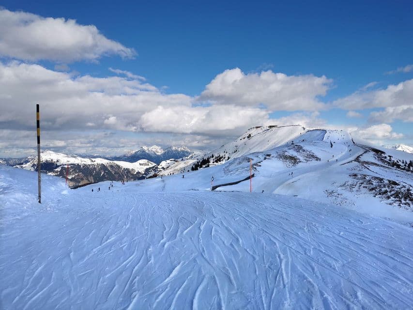 Une piste de ski damée avec des traces dans la neige mène vers des montagnes enneigées sous un ciel bleu avec des nuages blancs.