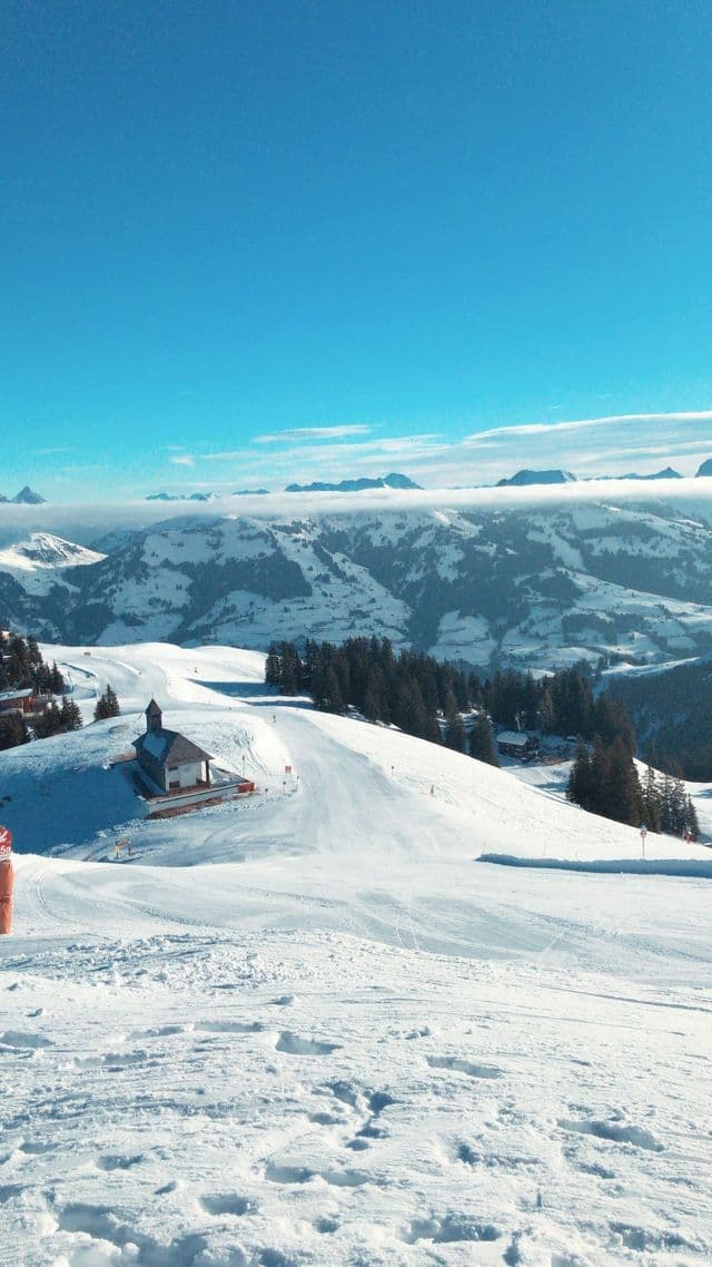 Une petite chapelle se trouve au bord d'une piste de ski, sur une chaîne de montagnes enneigée, sous un ciel bleu éclatant.