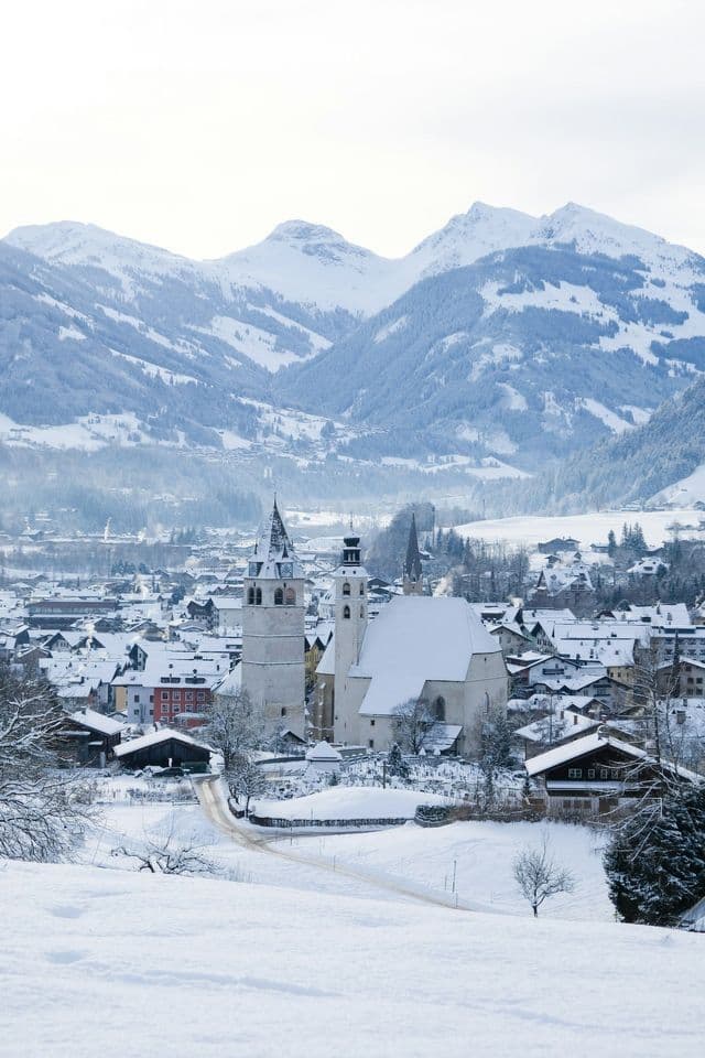Vogelperspektive auf eine verschneite Stadt mit zentraler Kirche, eingebettet in einem Tal, umgeben von schneebedeckten Bergen.