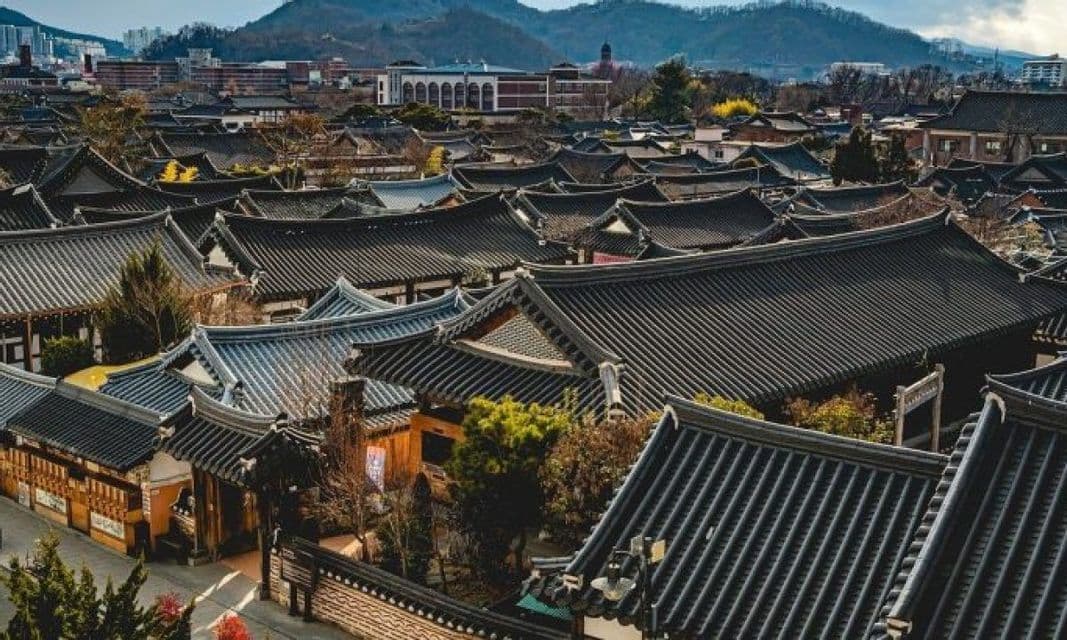 Veduta dall'alto di un villaggio tradizionale con tetti di tegole scure, con una città moderna e montagne sullo sfondo.