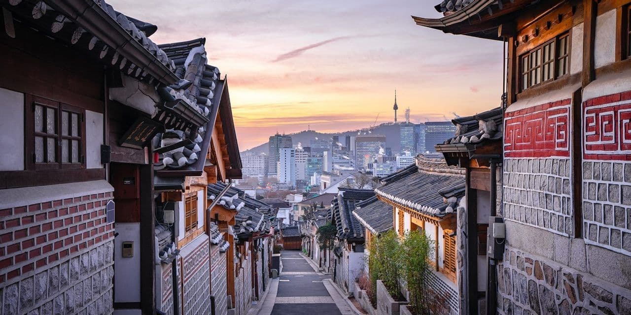 Una strada stretta in un tradizionale villaggio coreano con case dai tetti in tegole, che domina lo skyline moderno di una città con una torre al tramonto.