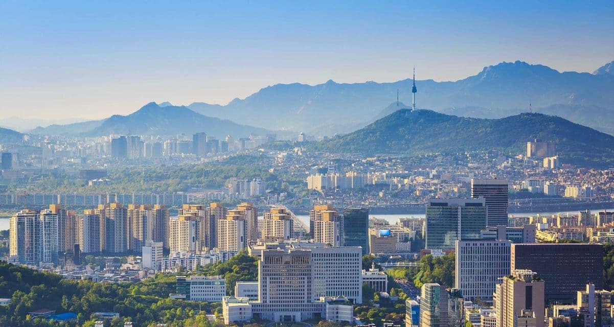 Veduta aerea di una vasta città con grattacieli ed edifici residenziali, con una torre prominente su una collina verde e montagne sullo sfondo.