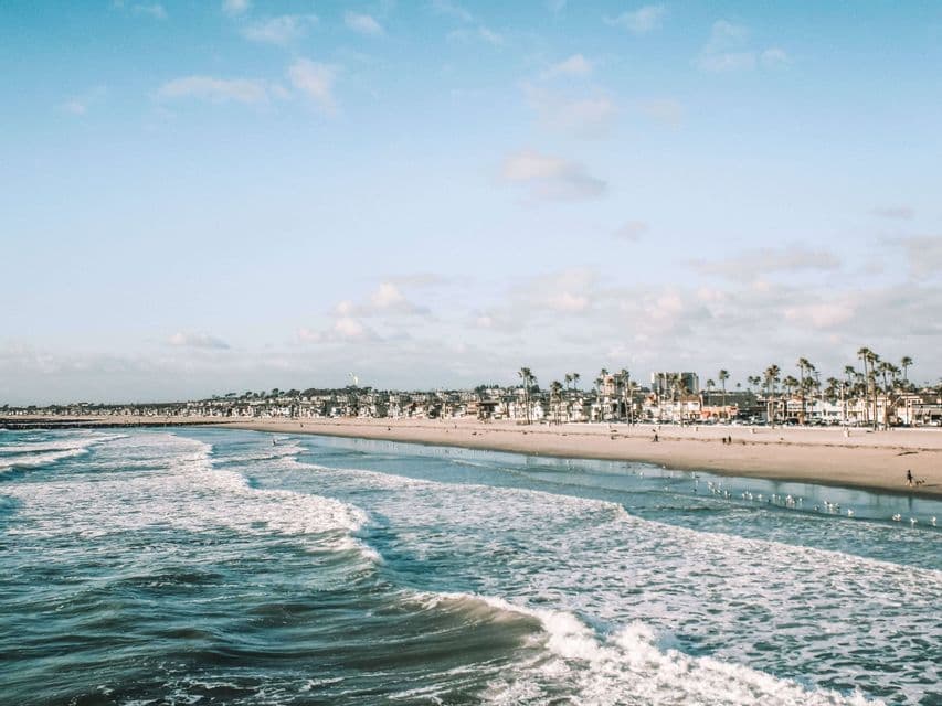 Les vagues de l'océan déferlent sur une vaste plage de sable, avec une ville côtière et des palmiers visibles à l'arrière-plan, sous un ciel bleu.