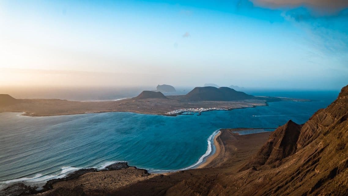 Una vista aerea di un'isola vulcanica con un piccolo villaggio costiero, circondata da acqua blu dell'oceano sotto un cielo sereno.