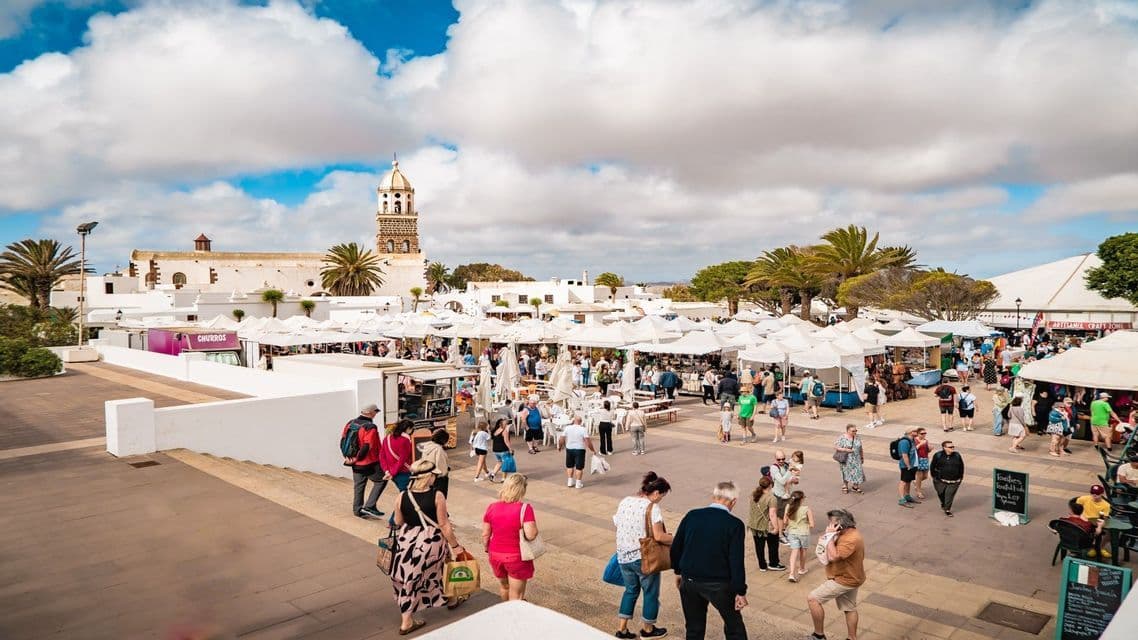 Una vista aérea de un bullicioso mercado al aire libre en una plaza del pueblo con puestos blancos, con una torre de iglesia al fondo bajo un cielo nublado.