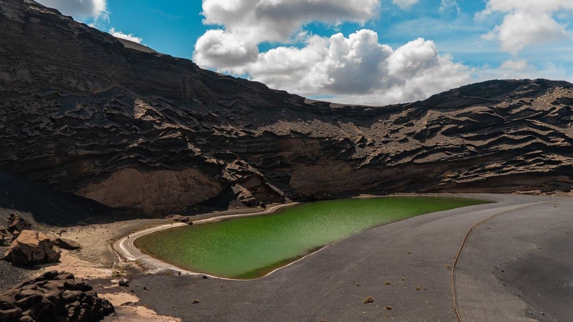 Un lago de un verde brillante, enclavado en la base de un oscuro y rocoso cráter volcánico bajo un cielo azul parcialmente nublado.