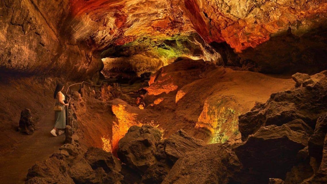 Una donna si trova su un sentiero all'interno di una vasta grotta rocciosa illuminata da calde luci arancioni e gialle.