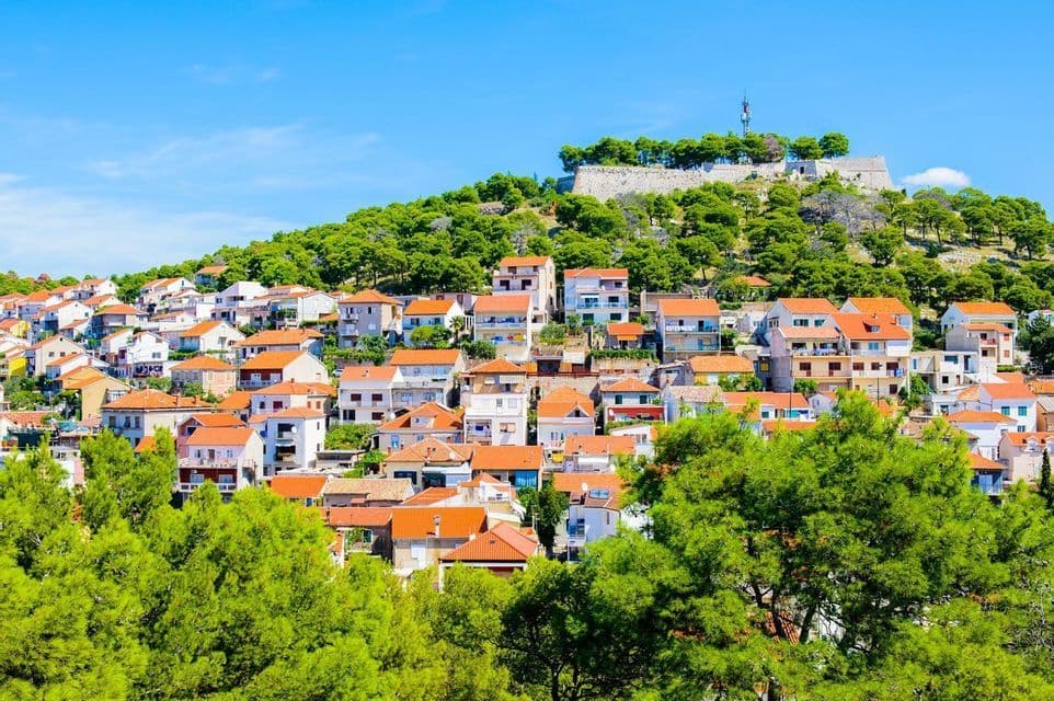 Ein Dorf mit weißen Gebäuden und roten Ziegeldächern, auf einem grünen Hügel erbaut, gekrönt von einer Steinfestung unter klarem Himmel.