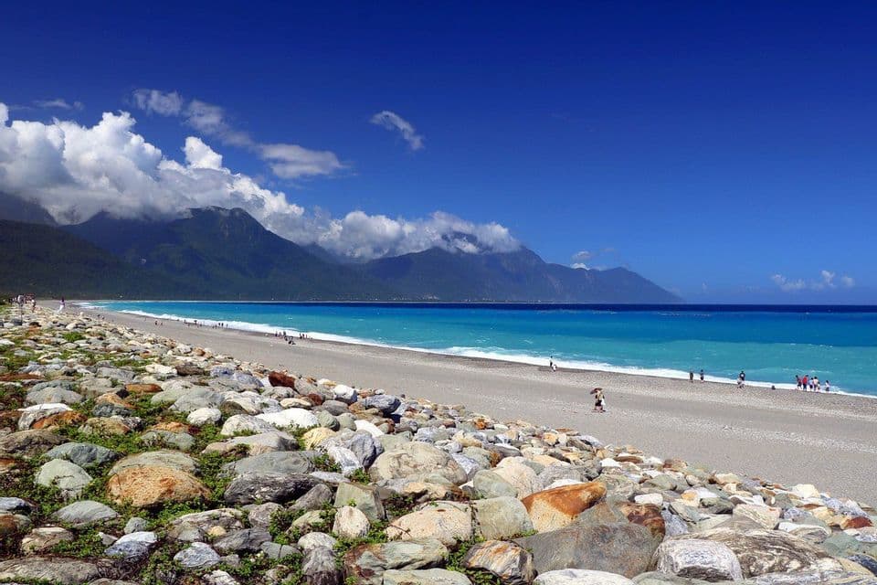 Una vista da una costa rocciosa, con una lunga spiaggia di ciottoli che incontra acque turchesi, montagne verdi sullo sfondo e un cielo azzurro.