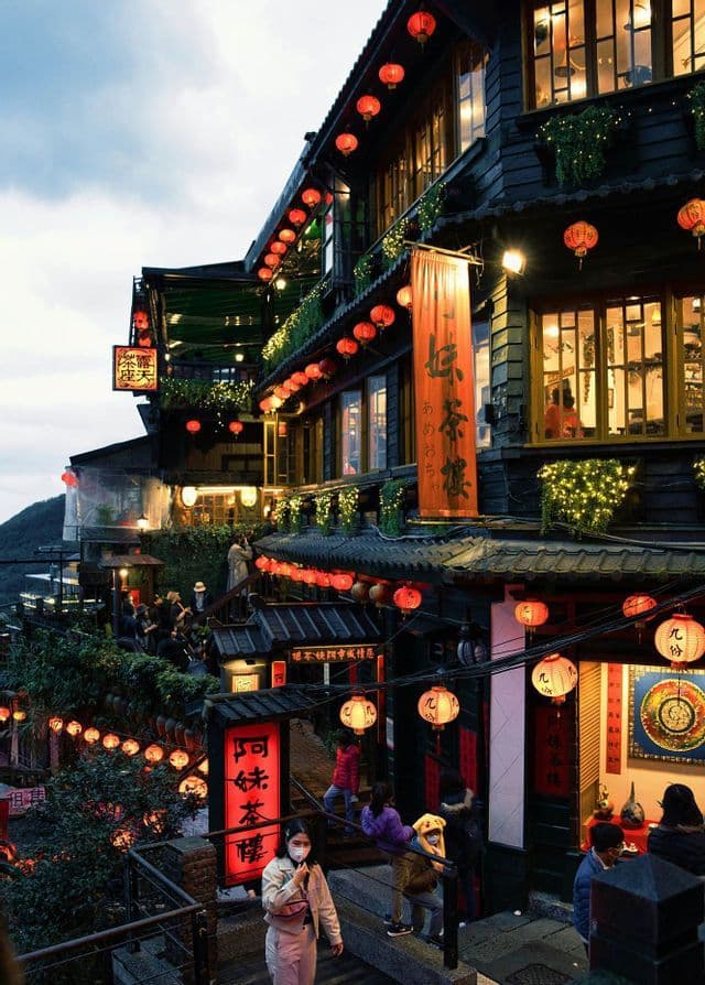 Al crepuscolo, le persone percorrono le scale tra edifici tradizionali collinari, illuminati da numerose lanterne di carta rosse e luminose.