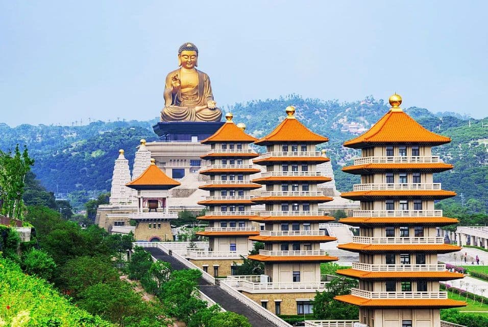 Una grande statua dorata di Buddha si erge in cima a un edificio, dominando diverse pagode con tetti arancioni su una lussureggiante collina verde.