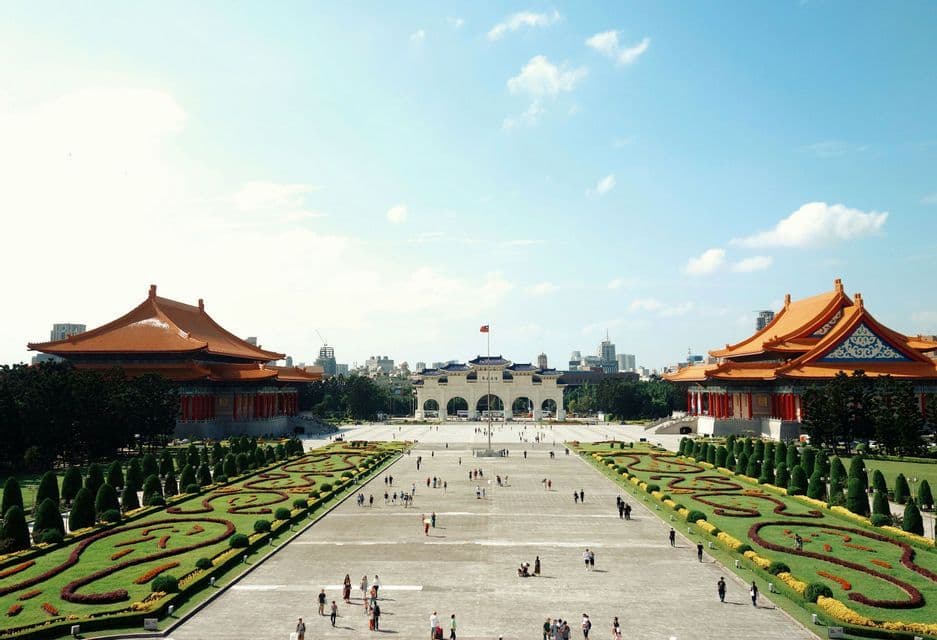 Una veduta ampia di una grande piazza pubblica affiancata da edifici tradizionali asiatici con tetti arancioni e giardini curati, con persone che passeggiano.