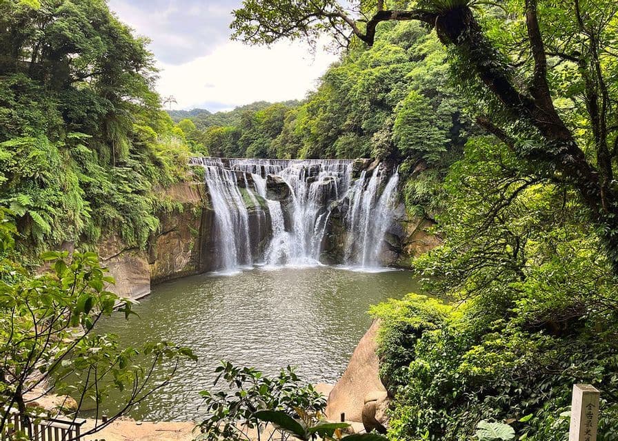 Una maestosa cascata si getta da una scogliera rocciosa in una pozza d'acqua, avvolta da una lussureggiante foresta verde sotto un cielo nuvoloso.