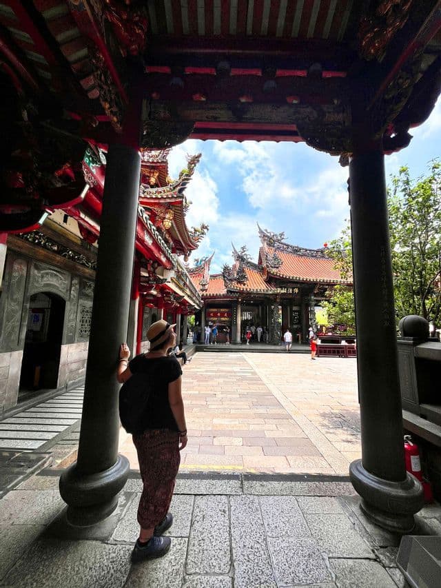 Una persona con cappello e zaino si trova sotto un arco rosso finemente decorato, osservando il cortile di un tradizionale tempio asiatico.