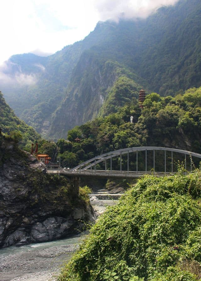 Un ponte ad arco attraversa una gola fluviale, circondata da montagne lussureggianti e verdi, con un tempio e una statua visibili sul pendio.