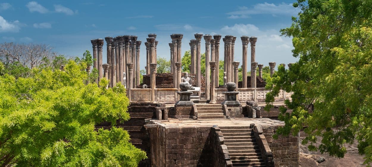 Les ruines d'un ancien temple avec des piliers de pierre et des statues sont encadrées par de luxuriants arbres verts sous un ciel bleu.