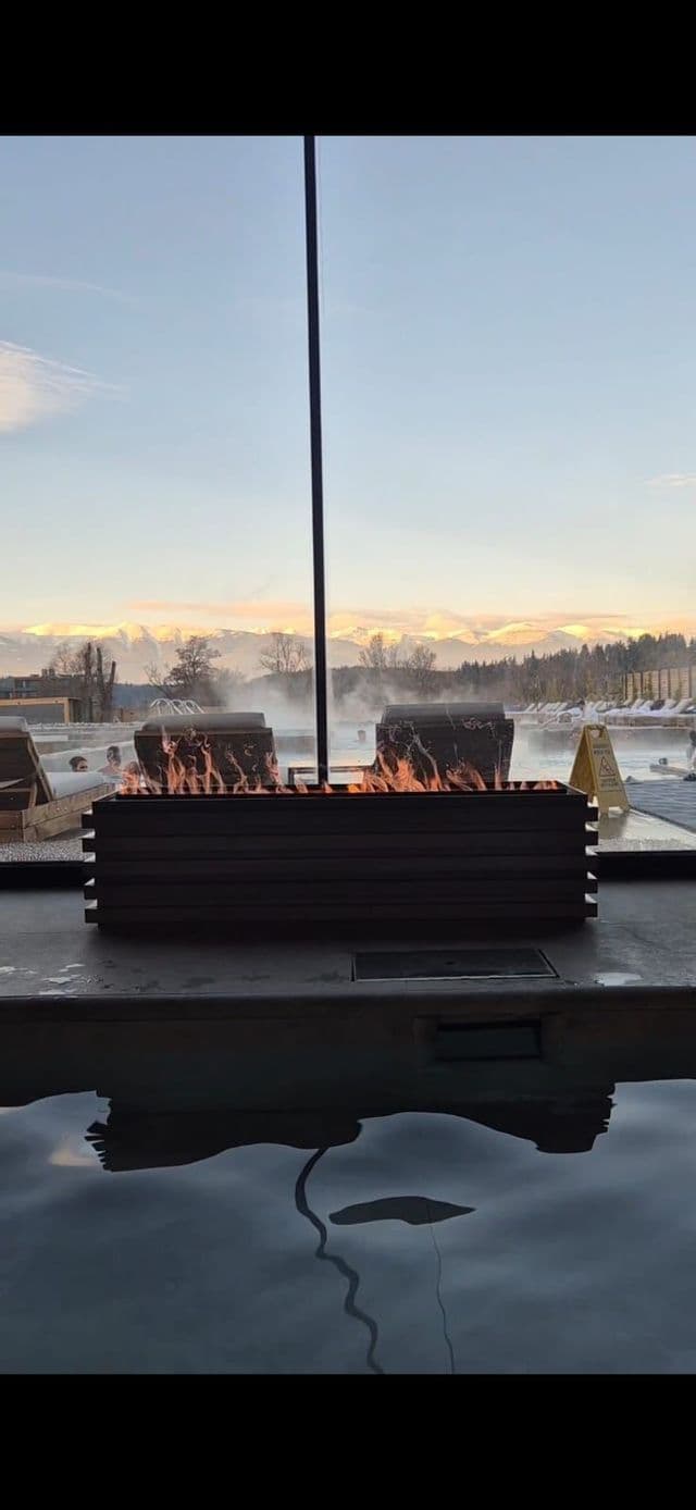 Eine Feuerstelle brennt neben einem dampfenden Thermal-Außenbecken mit Badenden und Fernblick auf schneebedeckte Berge.