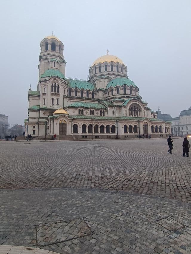 Une grande cathédrale avec des dômes dorés et des toits verts se dresse sur une vaste place pavée par un jour couvert.