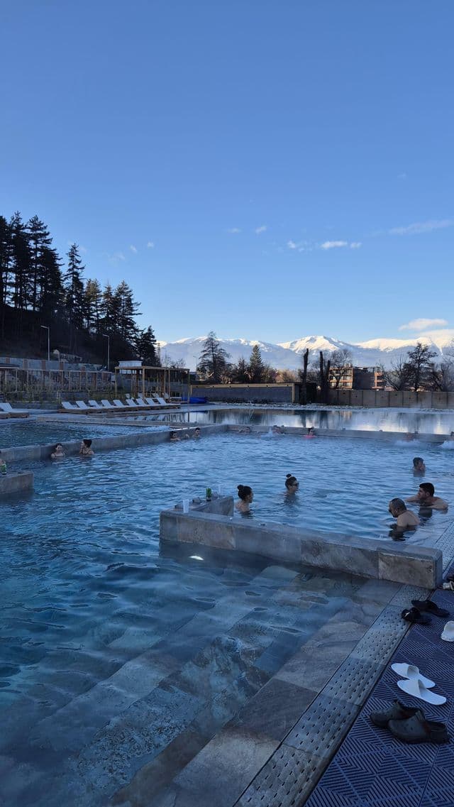 Un groupe WeRoad se détend dans une piscine extérieure fumante avec vue sur des montagnes enneigées sous un ciel dégagé.