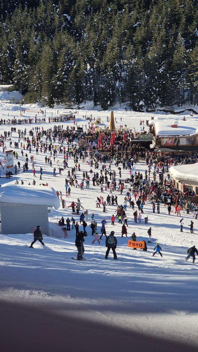 Vue plongeante d'une piste de ski bondée avec de nombreuses personnes skiant et socialisant près des chalets au pied d'une forêt de pins.
