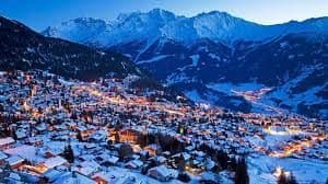 Un village de montagne enneigé, illuminé par des lumières au crépuscule, niché dans une vallée avec de grands sommets en arrière-plan.