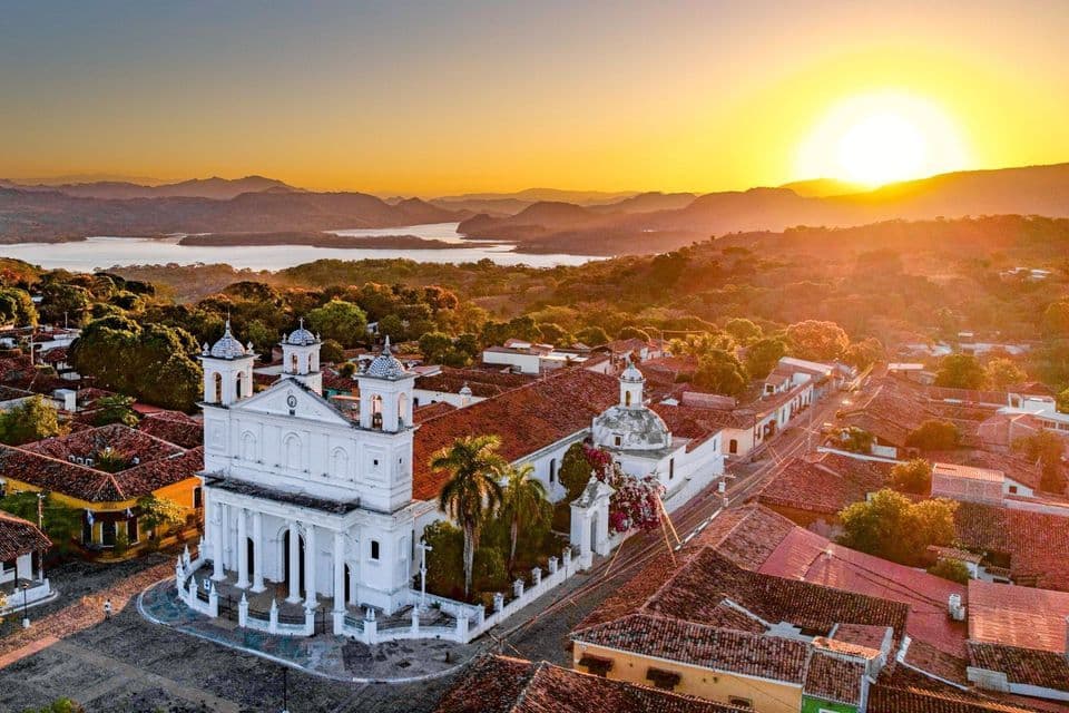Luftaufnahme einer weißen Kolonialkirche in einer Stadt mit roten Ziegeldächern bei Sonnenuntergang, mit einem See und Hügeln in der Ferne.