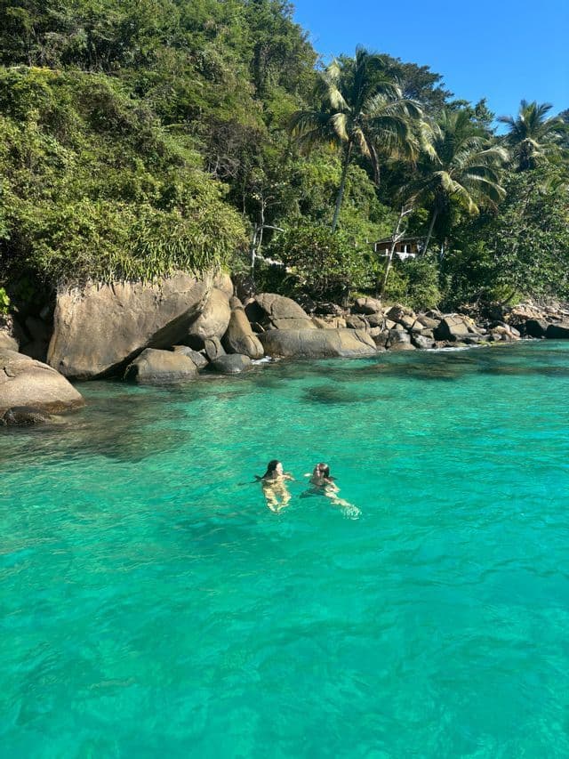 Dos personas de un viaje en grupo de WeRoad nadando en aguas turquesas cristalinas junto a una costa rocosa cubierta de exuberante vegetación.