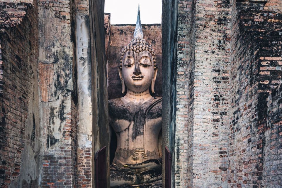 Una grande statua di Buddha in pietra, dall'espressione serena, si trova incorniciata tra due muri di mattoni logori.