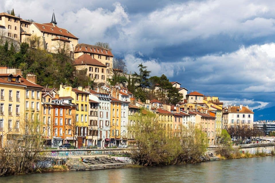 Una fila di edifici colorati si erge sulla riva di un fiume, ai piedi di una collina verdeggiante, sotto un cielo parzialmente nuvoloso.