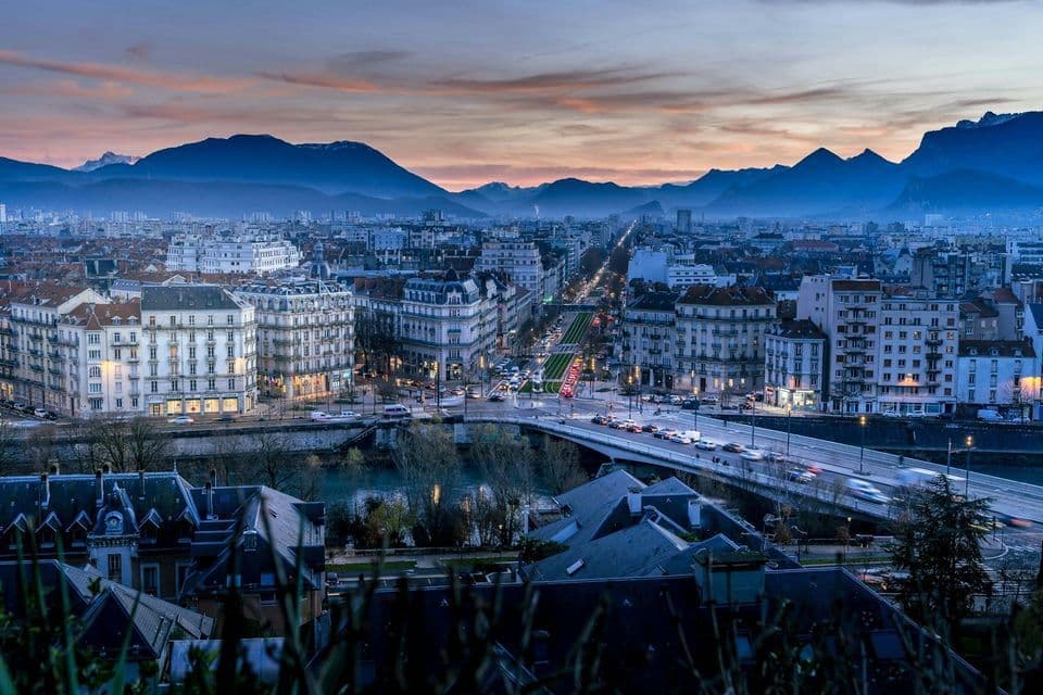 Vista elevada de un paisaje urbano con edificios iluminados y tráfico en un puente, con una cordillera al atardecer de fondo.