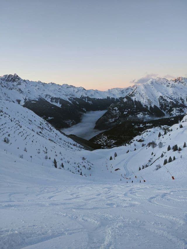 A snow-covered mountain slope with ski tracks overlooks a valley filled with a dense layer of clouds at dusk.