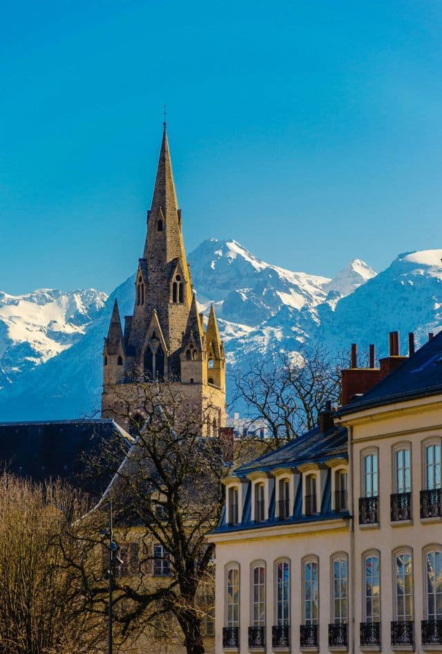 Una guglia di chiesa in pietra svetta sui tetti della città, con uno sfondo di montagne innevate sotto un cielo azzurro chiaro.