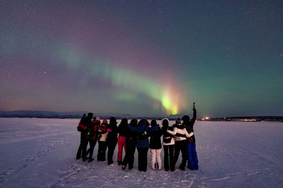 A WeRoad group trip stands together in a snowy landscape at night, watching the aurora borealis under a starry sky.