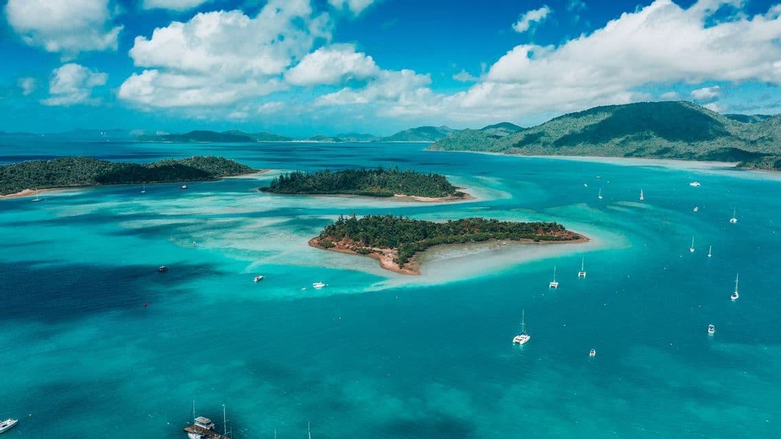 An aerial view of sailboats and catamarans floating in turquoise water surrounding small, forested islands under a blue sky with clouds.
