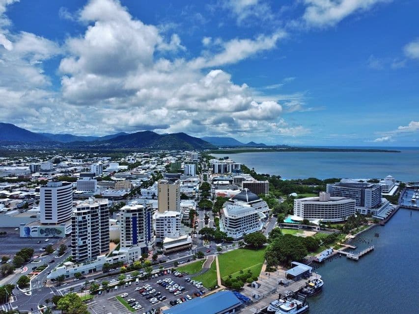 Une vue aérienne d'une ville côtière avec des bâtiments modernes, un port et des montagnes vertes en arrière-plan sous un ciel bleu, partiellement nuageux.