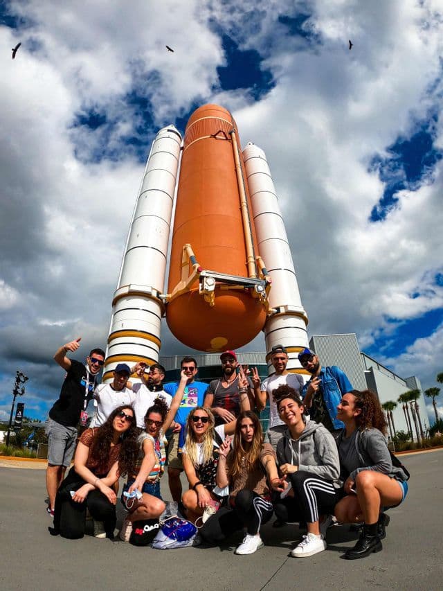 Un gruppo WeRoad in viaggio posa per una foto di fronte a una replica di un razzo space shuttle sotto un cielo nuvoloso.