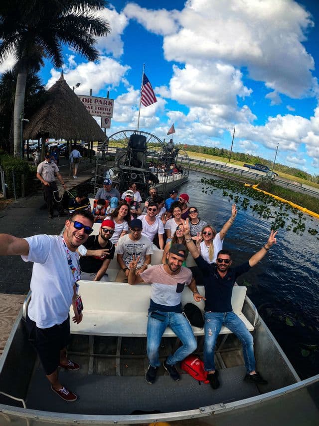 Eine WeRoad-Gruppe lächelt und posiert für ein Selfie auf einem Airboat auf dem Wasser unter einem blauen, bewölkten Himmel.