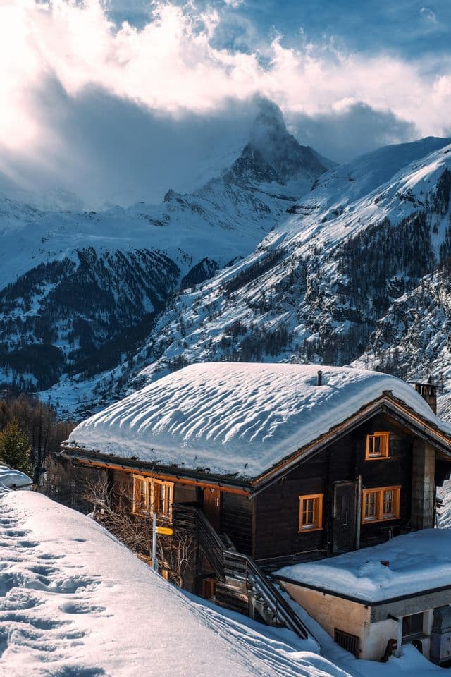 Un chalet en bois au toit enneigé est perché à flanc de montagne, avec un sommet spectaculaire visible derrière lui à travers les nuages.