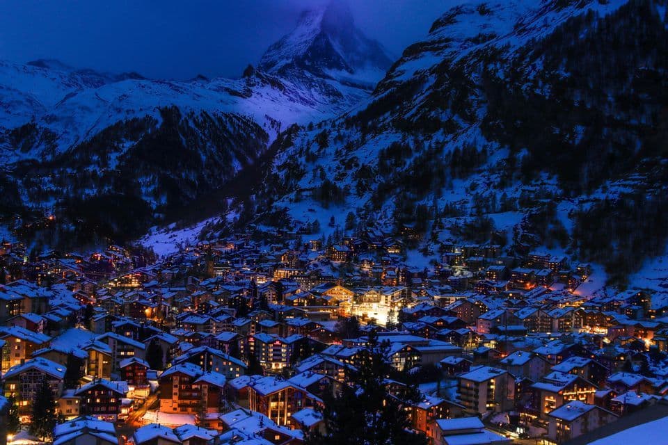 An illuminated village with snow-covered roofs sits in a mountain valley at dusk, with a towering, snow-capped peak in the background.