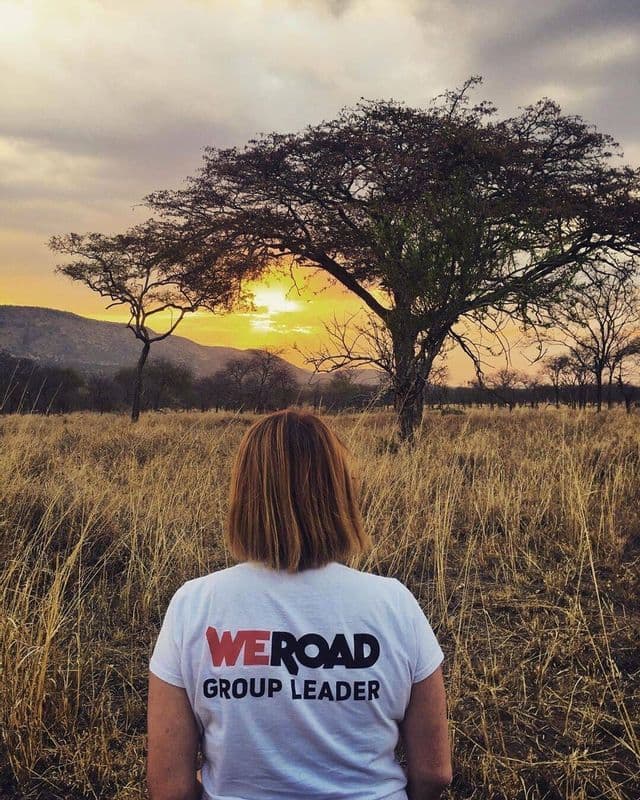Ein WeRoad Gruppenleiter, von hinten, steht in einem Savannenfeld und beobachtet den Sonnenuntergang hinter einem großen Baum.