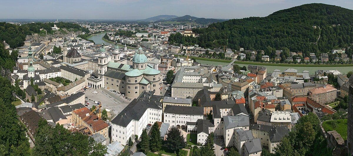 Una vista panoramica dall'alto di una città storica con una cattedrale a cupola prominente accanto a un fiume e una collina boscosa.