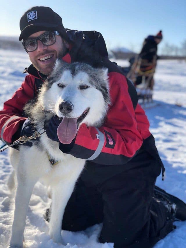 Ein lächelnder Mann mit roter Jacke und Sonnenbrille kniet im Schnee, um einen Husky-Hund mit heraushängender Zunge zu umarmen.