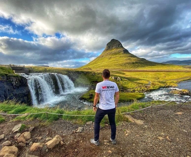 A WeRoad group leader seen from behind, looking at a waterfall flowing next to a large green mountain under a cloudy sky.