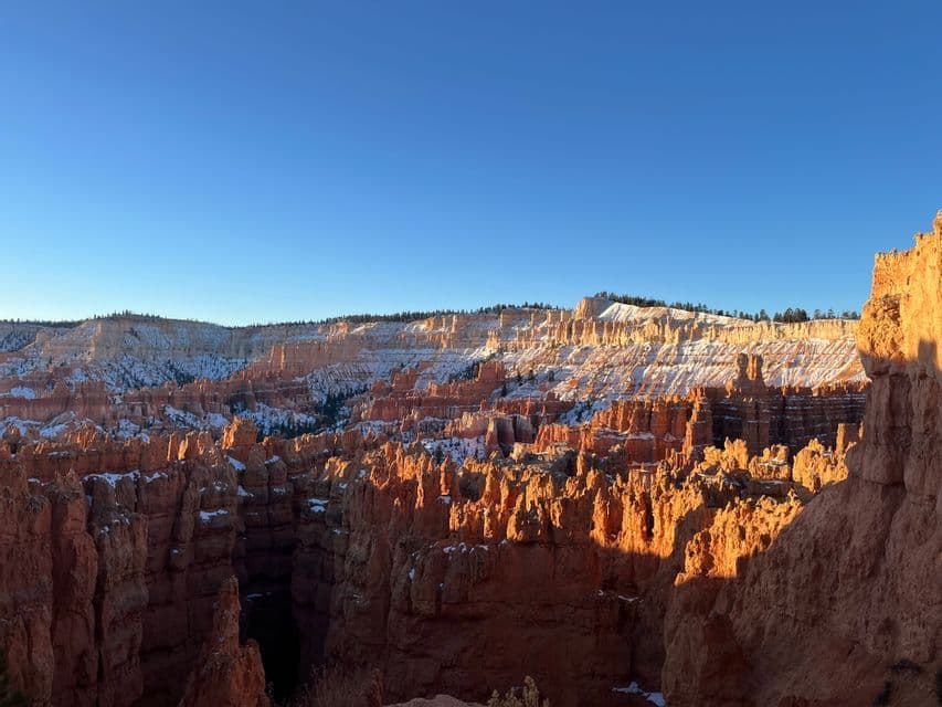 Un'ampia vista di un canyon con formazioni rocciose rosse spolverate di neve, illuminate dalla luce del sole mattutino sotto un cielo azzurro chiaro.