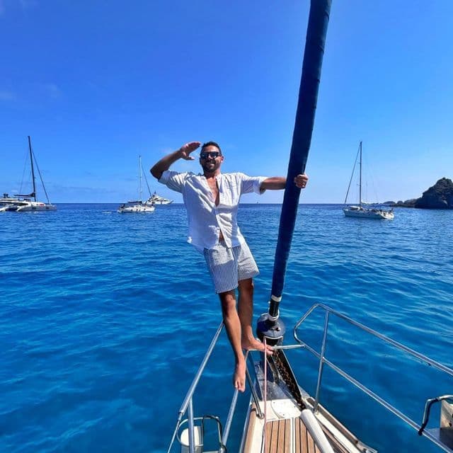 Ein lächelnder Mann mit Sonnenbrille steht am Bug eines Segelboots und hält den Mast, während es auf strahlend blauem Meer, umgeben von anderen Booten, segelt.